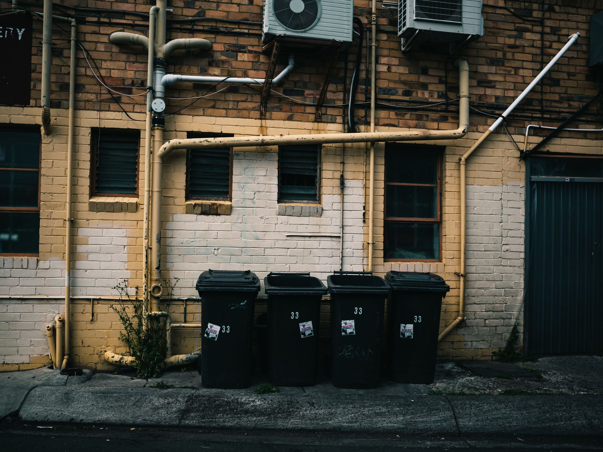 Back alley scene with trash bins and air conditioners on a brick wall.